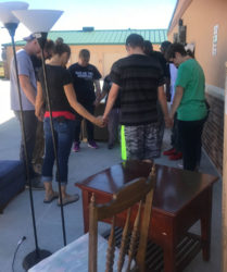 Group of people holding hands in a circle on a sunny patio, surrounded by various furniture items and moving boxes.