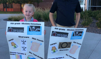Two children holding donation posters for a 4th-grade class mission, standing in front of a school building.