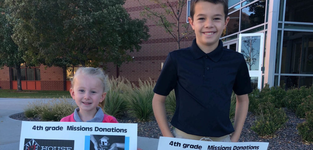 Two children holding donation posters for a 4th grade mission project outside a school building.