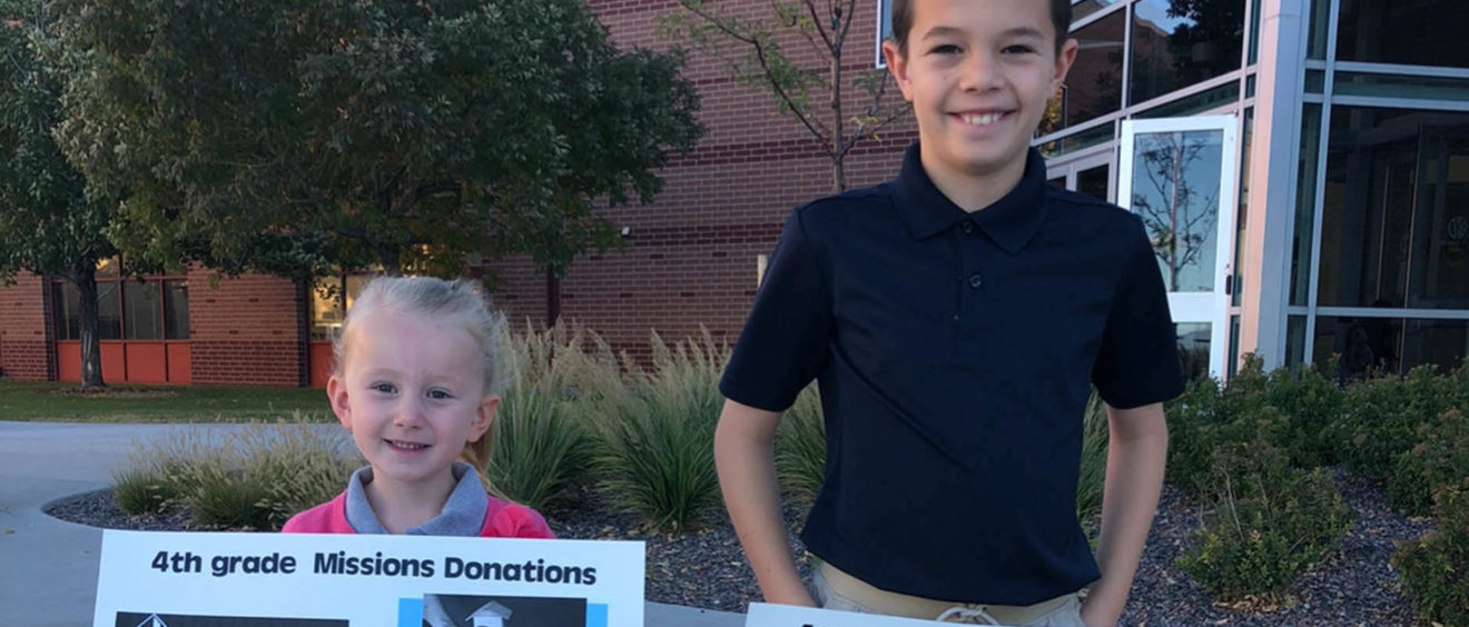 Two children holding donation posters for a 4th grade mission project outside a school building.