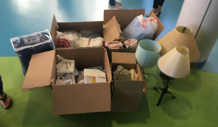 Cardboard boxes filled with various items like clothes and lamps on a green floor, while two people stand nearby.