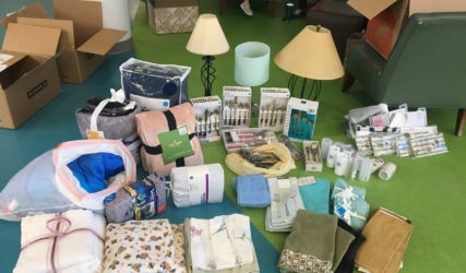 Assorted household items and moving boxes spread out on a green floor, including lamps, linens, and kitchenware.