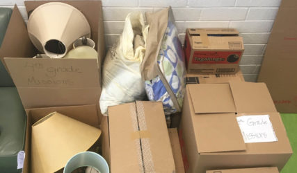 Pile of assorted unpacked and labeled cardboard boxes against a white brick wall, containing various items and school materials.