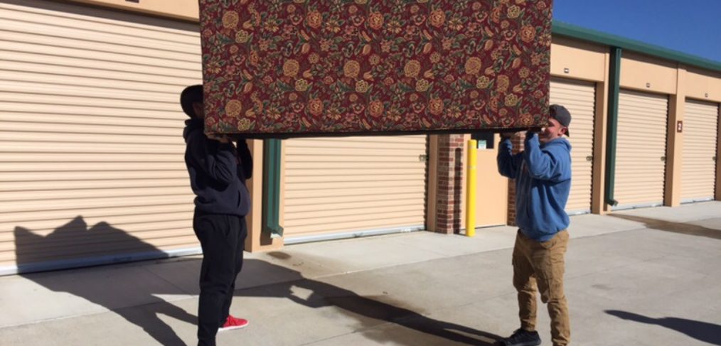 Two people carrying a large, ornate rug outside a storage facility, under a clear blue sky.