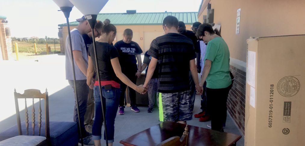 Group of people holding hands and praying outdoors next to stacked boxes and furniture.