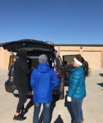 Three people, one wearing a blue jacket and two in black, stand at the open trunk of an suv in a parking lot.