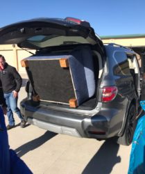 A large piece of furniture loaded into the trunk of an suv, with several people gathered around, under a clear sky.