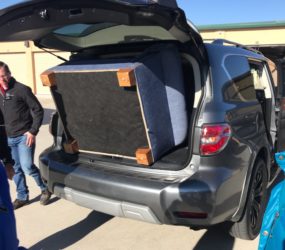 A large piece of furniture loaded into the trunk of an suv, with several people gathered around, under a clear sky.