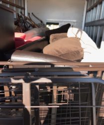 Storage unit interior with mixed items including a beige cushion on a black sofa, visible metal tools, furniture, and a collapsible black crate.