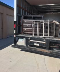 Flatbed truck loaded with a metal cage and a multi-drawer tool chest parked beside storage units under a clear sky.