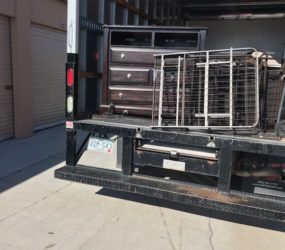 Flatbed truck loaded with a metal cage and a multi-drawer tool chest parked beside storage units under a clear sky.