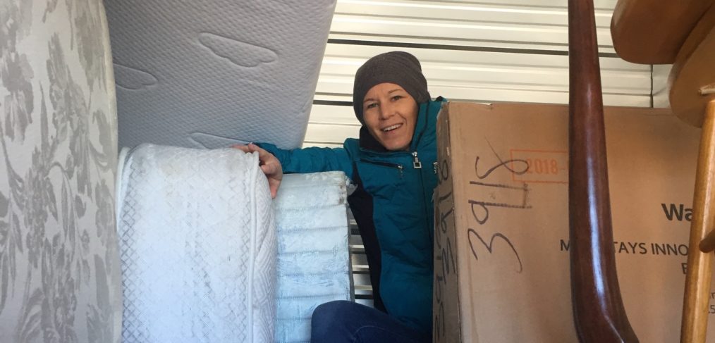 A woman in a blue jacket and beanie smiles while standing amidst stacked mattresses and boxes in a storage unit.