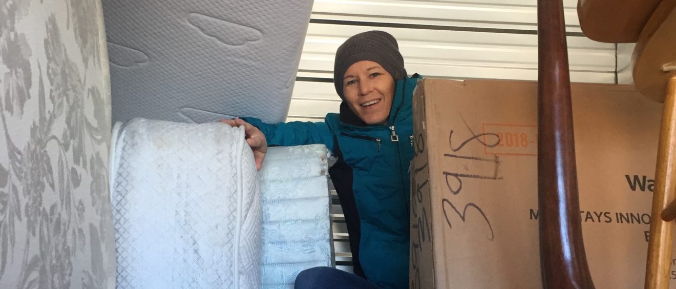 A woman in a blue jacket and beanie smiles while standing amidst stacked mattresses and boxes in a storage unit.