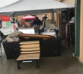 A person stands under a tent amidst snowfall, organizing various items and furniture outside a storage unit.