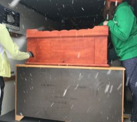 Two people loading a large wooden chest into a truck during a snowfall. one person is wearing a green jacket and the other is in an orange beanie.