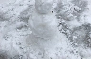 A small, roughly-built snowman standing on a snow-covered pavement with tire tracks in the background.