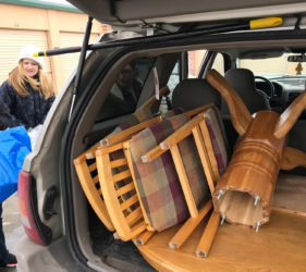 A woman smiling near an open car trunk filled with disassembled wooden furniture, including chairs and a table.