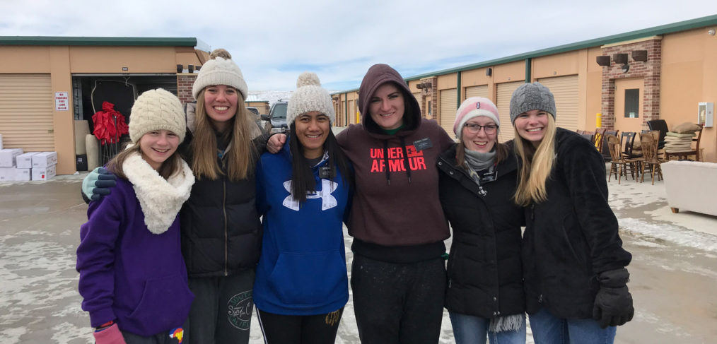 Six women smiling and standing together outdoors, wearing winter hats and coats, with a snowy background and buildings behind them.