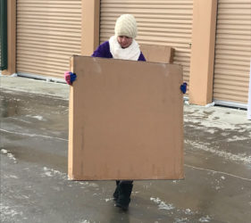 Person in winter clothing carrying a large cardboard sign in front of a building with storage units.