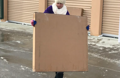 Person in winter clothing carrying a large cardboard sign in front of a building with storage units.