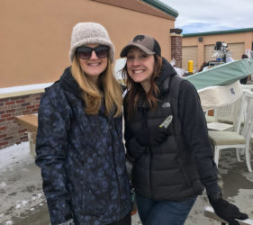 Two women in winter clothing smiling together outdoors with a snow-covered ground and a beige building in the background.