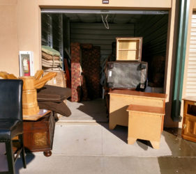 A storage unit open with various household items, including furniture and boxes, scattered in front and inside the unit.