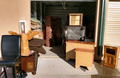 A storage unit open with various household items, including furniture and boxes, scattered in front and inside the unit.