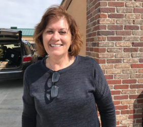 A woman smiles standing outside by a brick wall, wearing a blue sweater and sunglasses hanging from her collar.