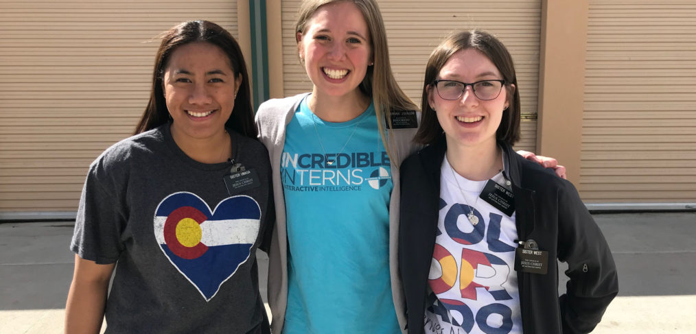 Three women smiling at the camera, standing together outside a building. they are wearing different graphic t-shirts.