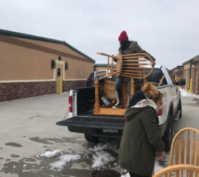 A person loads wooden chairs into the bed of a pickup truck in a parking lot, with another person assisting by handing over a chair.