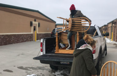 A person loads wooden chairs into the bed of a pickup truck in a parking lot, with another person assisting by handing over a chair.