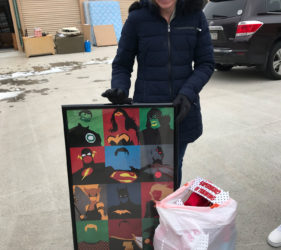 Woman smiling, holding a framed artwork featuring colorful superhero masks, standing in a parking lot with a bag of gifts.