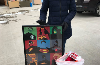 Woman smiling, holding a framed artwork featuring colorful superhero masks, standing in a parking lot with a bag of gifts.