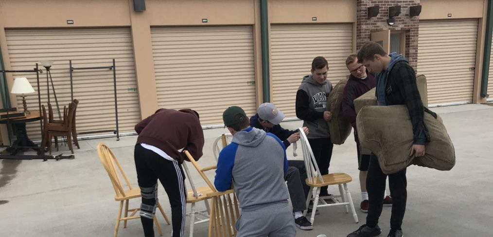 Group of people organizing chairs and items outside a storage unit under a cloudy sky.