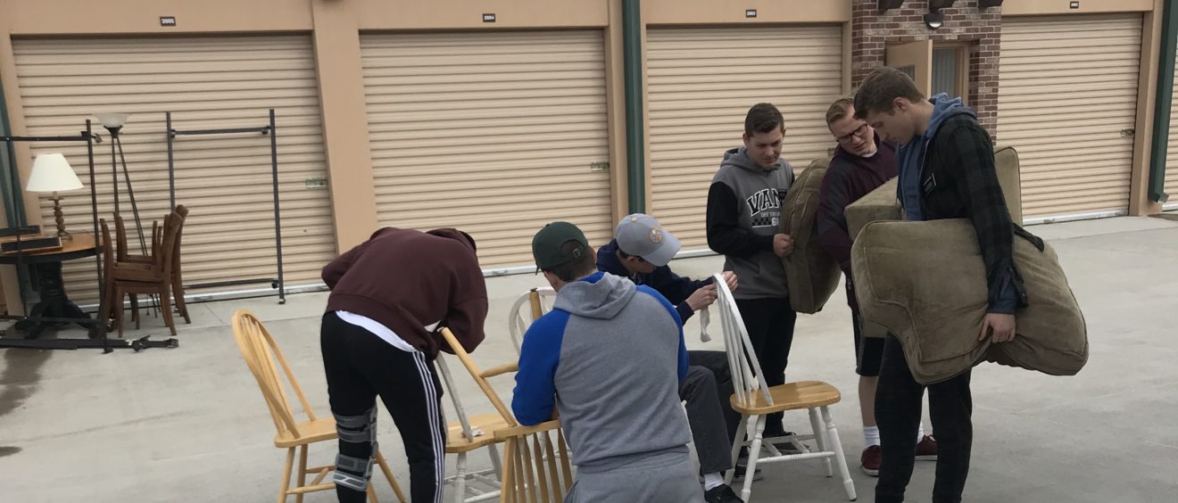 Group of people organizing chairs and items outside a storage unit under a cloudy sky.