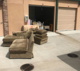 A collection of discarded furniture including a sofa, chairs, and cushions outside a storage facility under a cloudy sky.