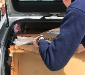 A person loading a cardboard box into the back of a car, with another box and a pink bag already inside the trunk.