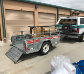 A u-haul trailer attached to a blue and white pickup truck, parked in front of storage units with an open trailer ramp and various items loaded inside.