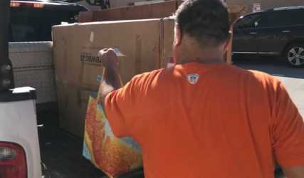 A man in an orange shirt unloading large cardboard boxes from the back of a pickup truck in a sunny parking lot.