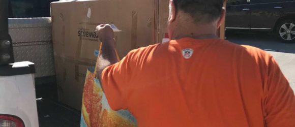 A man in an orange shirt unloading large cardboard boxes from the back of a pickup truck in a sunny parking lot.