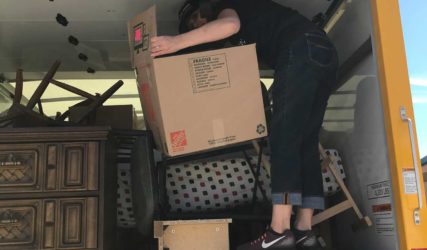 A person loading a large cardboard box into a packed moving truck, stepping on furniture to reach inside.