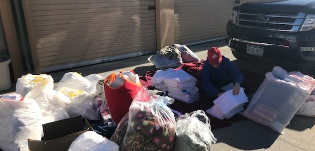 Person sorts through various items next to a parked car in front of an open storage unit under bright sunlight.