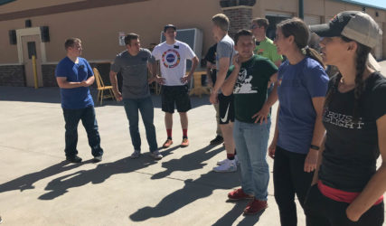 A group of people standing in a sunny outdoor area near a building, engaged in a conversation, with clear skies overhead.