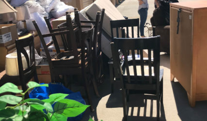 Two people standing in a storage area with discarded furniture and boxes under a clear sky.